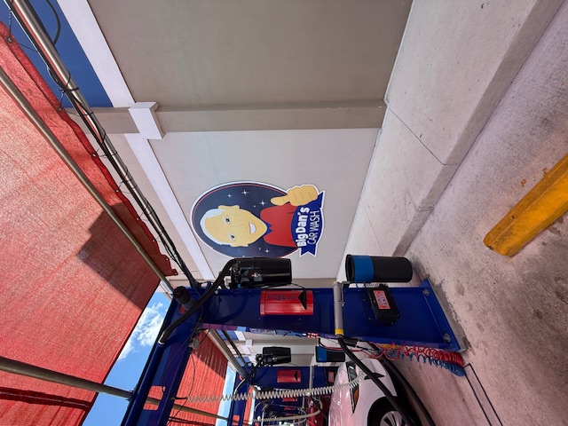 The Big Dan's Car Wash mascot logo painted on the exterior wall — a smiling silver-haired man in a red shirt giving a thumbs-up inside a starry circle, with the Big Dan's Car Wash banner across it