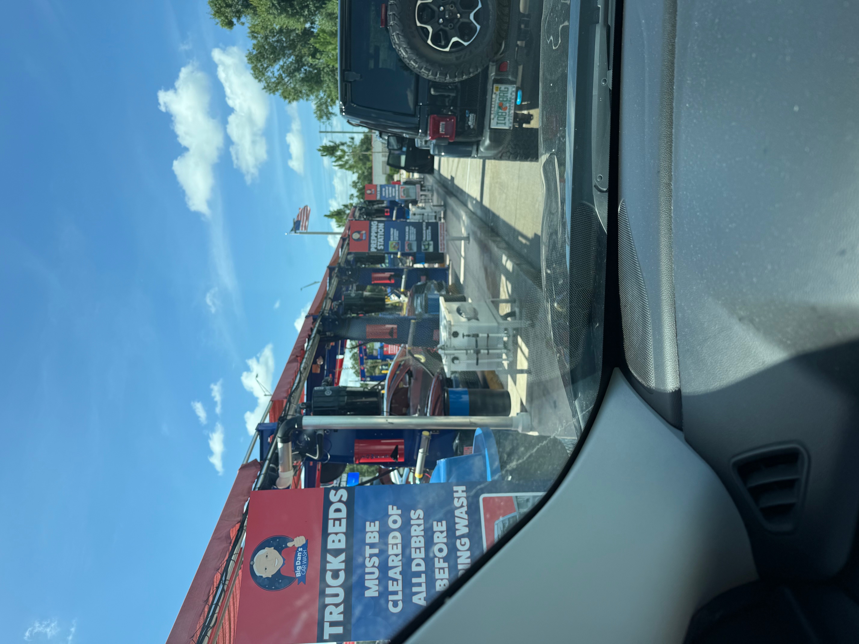 The view down Big Dan's Car Wash prep lane from a vehicle waiting in line — a Prepping Station sign and a Truck Beds Must Be Cleared Of All Debris sign on the red canopy, with prep equipment lining the covered bays