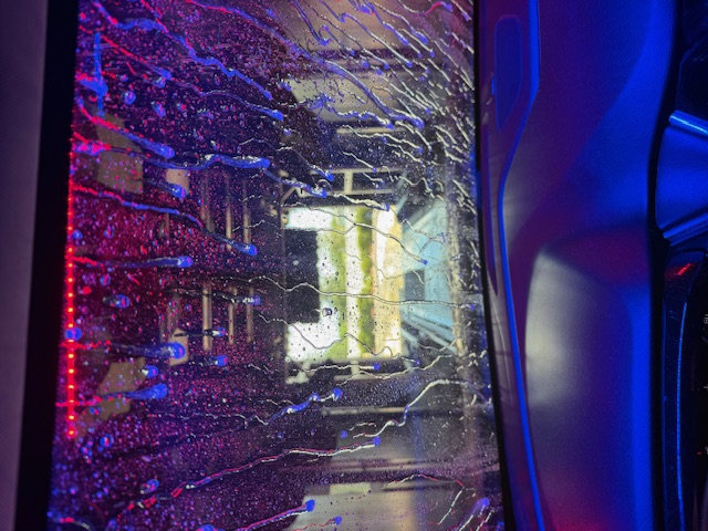 Inside the Big Dan's Car Wash tunnel during a wash — dripping cloth wash mitters and water streaking the windshield under red, blue, and pink LED lighting, with the tunnel exit visible ahead