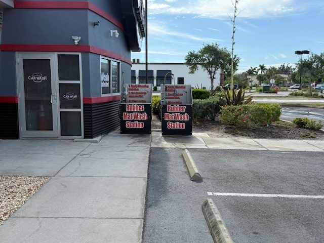 Two Rubber Mat Wash Station signs outside River Club Car Wash, each marking a dedicated bay with cleaning instructions posted above