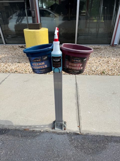 Free towel and window cleaner station at River Club Car Wash — a blue clean towels bucket and a burgundy dirty towels return bucket flanking a mounted window cleaner spray bottle