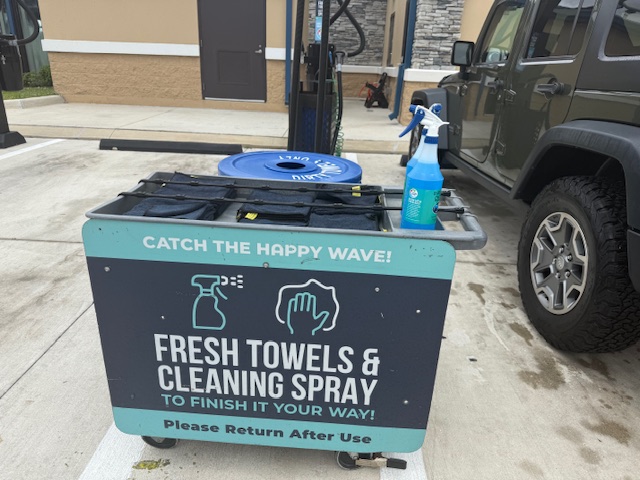 Close-up of the Tidal Wave Auto Spa towel cart — branded 'Catch the Happy Wave! Fresh Towels and Cleaning Spray' with a spray bottle of glass cleaner and clean and dirty towel sections